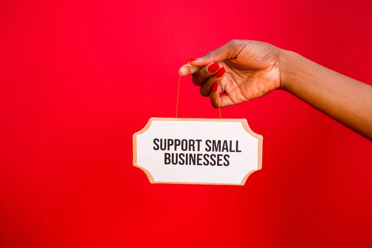 A hand holds a sign that reads Support Small Businesses against a vibrant red background.